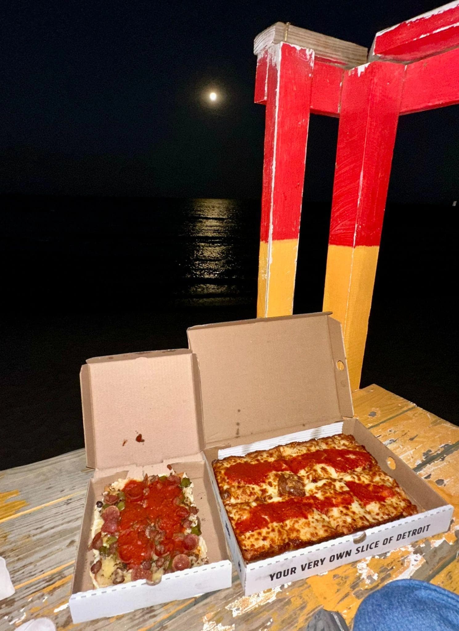 Two open pizza boxes beside the water at night under a full moon